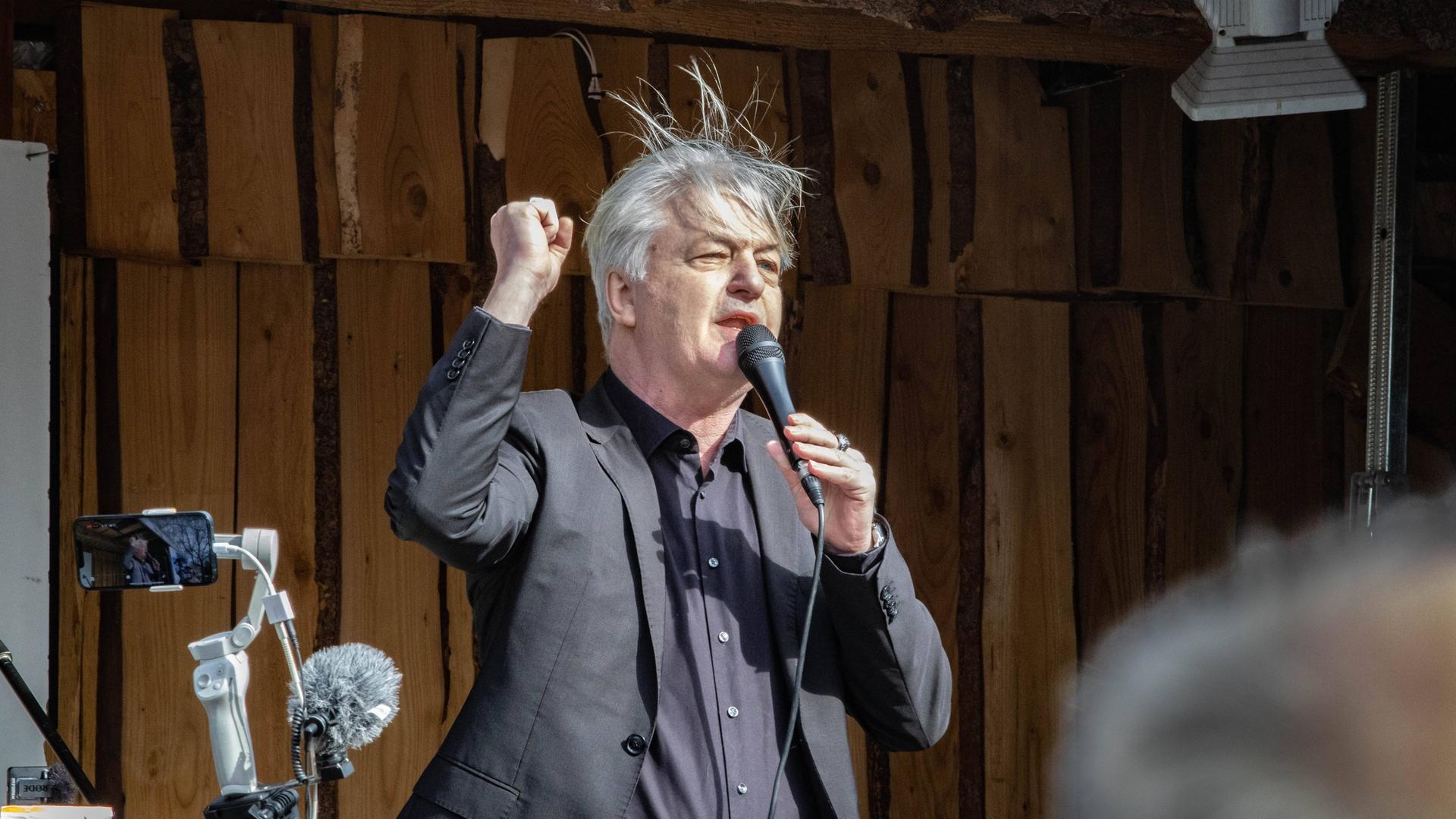 Ein Mann mit wirrem Haar reckt seine rechte Faust in die Luft. Er spricht in eine Mikrofon und steht offenbar auf einer Bühne.