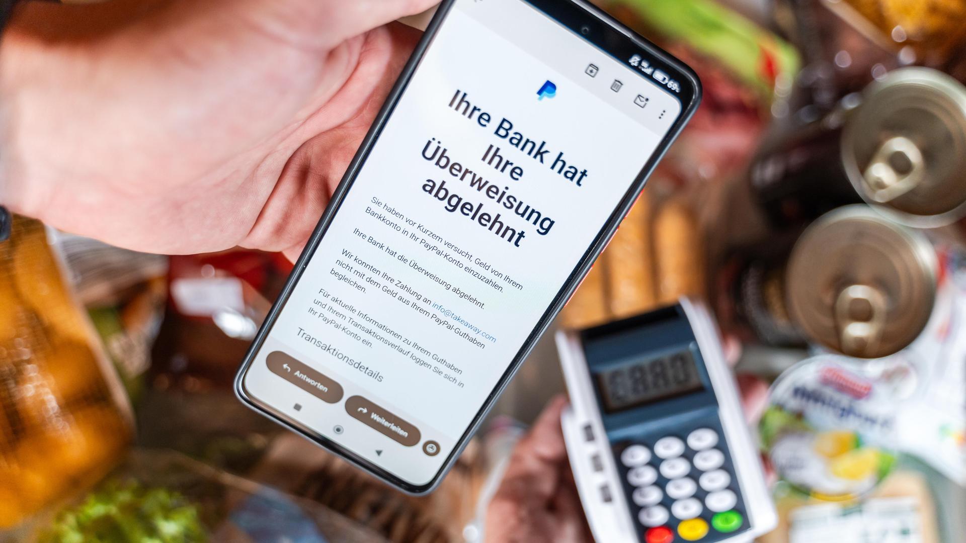 Ein Smartphone zeigt eine Paypal-Mitteilung über eine abgelehnte Überweisung, darunter ist ein Kartenlesegerät zu sehen. (Symbolbild) Ein Smartphone zeigt eine Paypal-Mitteilung über eine abgelehnte Überweisung, darunter ist ein Kartenlesegerät zu sehen. (Symbolbild)