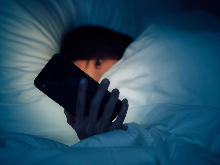 Zu sehen ist eine Frau, eingehüllt in eine dicke Bettdecke. Sie blickt auf ein leuchtendes Smartphone, dass ihr besorgtes Gesicht im Dunkeln erhellt.