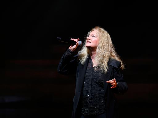Nicole steht bei ihrer Jubiläumstournee in der Meistersingerhalle Nürnberg mit einem Mikrofon posierend vor schwarzem Hintergrund auf der Bühne.