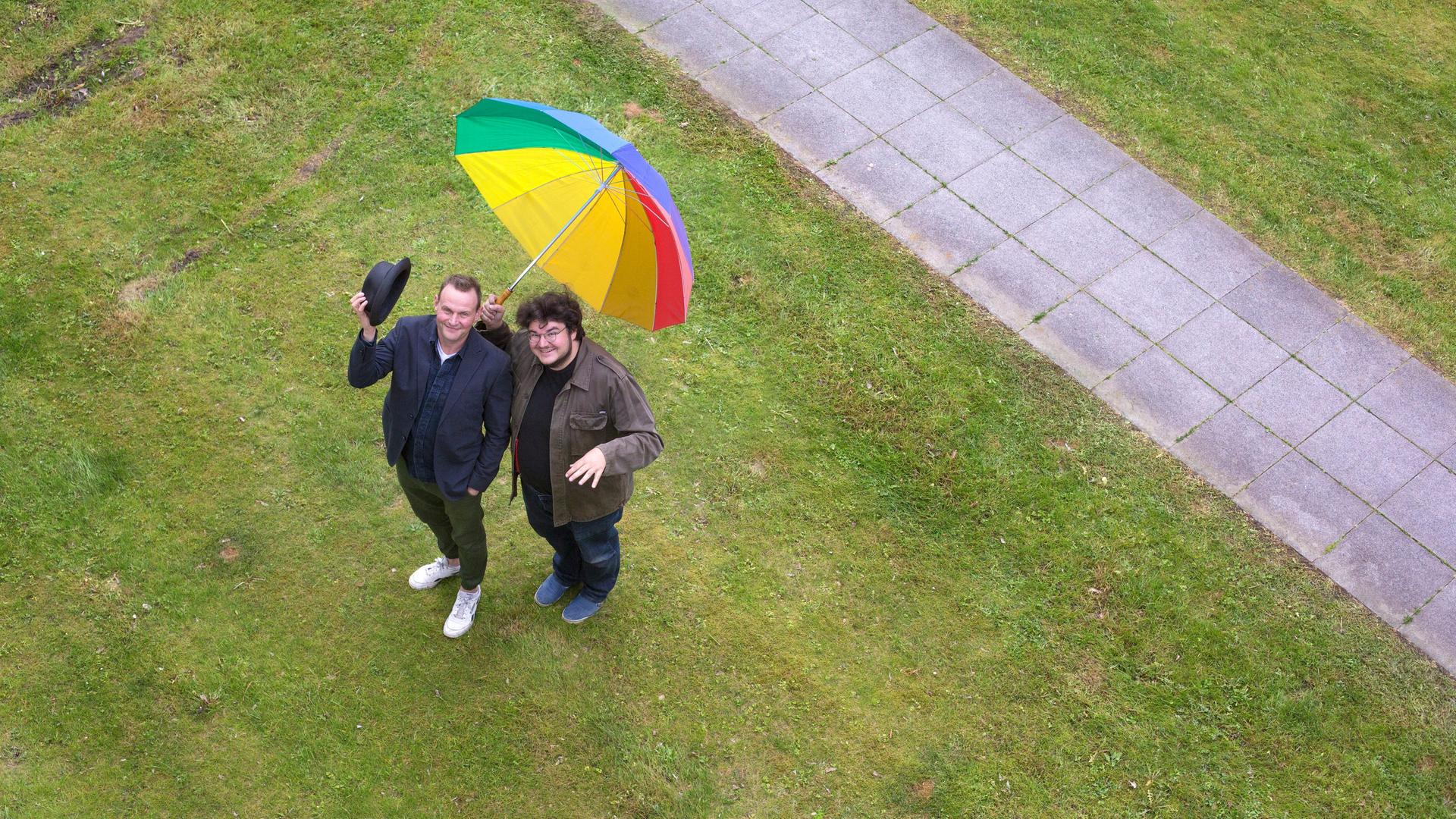 Devid Striesow und Axel Ranisch auf einer Wiese mit regenbogenfarbenen Regenschirm. Das Bild ist aus der Vogelperspektive fotografiert, beide schauen nach oben.