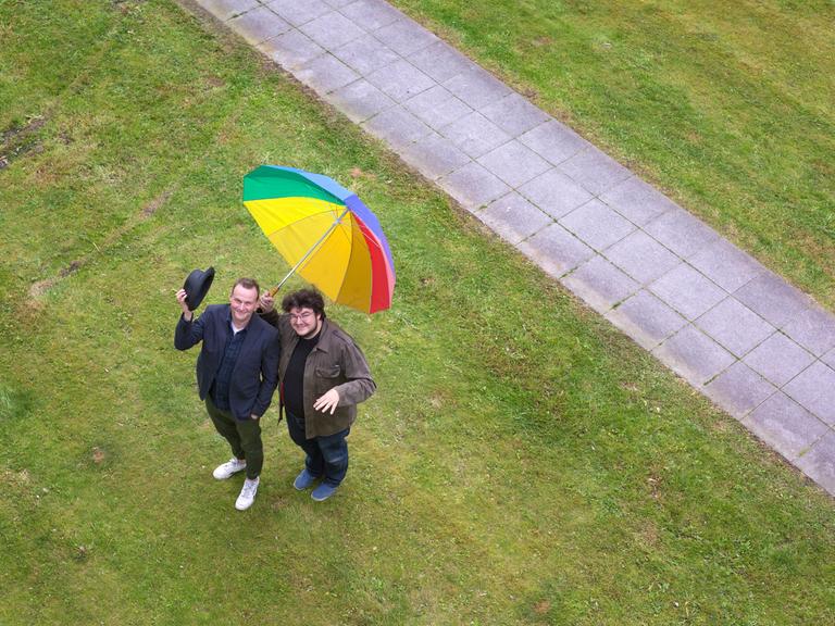 Devid Striesow und Axel Ranisch auf einer Wiese mit regenbogenfarbenen Regenschirm. Das Bild ist aus der Vogelperspektive fotografiert, beide schauen nach oben.