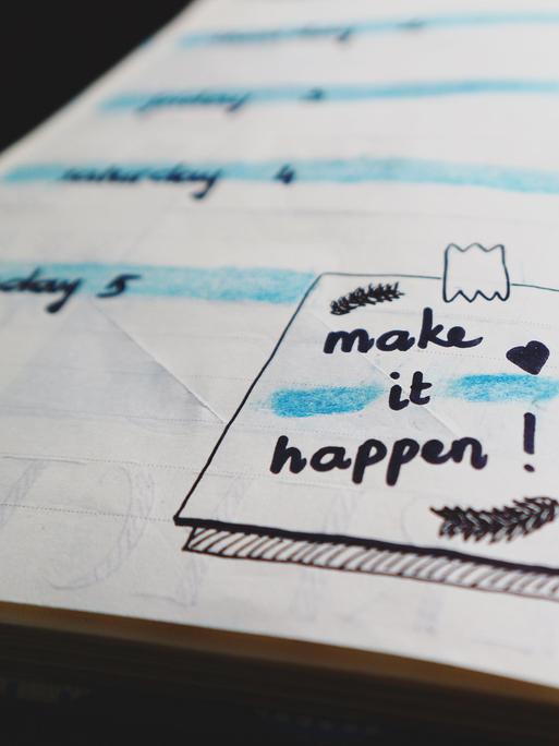 Ein aufgeschlagener Kalender enthält eine kleine Zeichung, die den Schriftzug "Make it happen" trägt. 