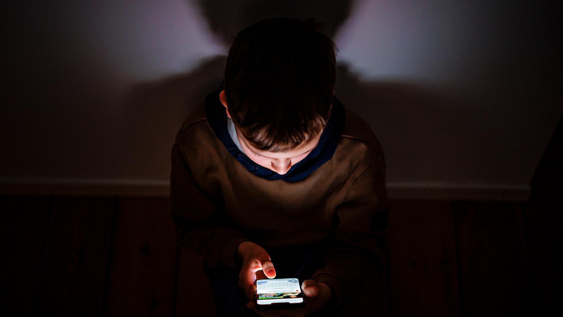 Symbolfoto zum Thema Kinder nutzen Social Media: Ein Junge sitzt in einem dunklen Raum, sein Gesicht wird von einem Smartphone beleuchtet.
