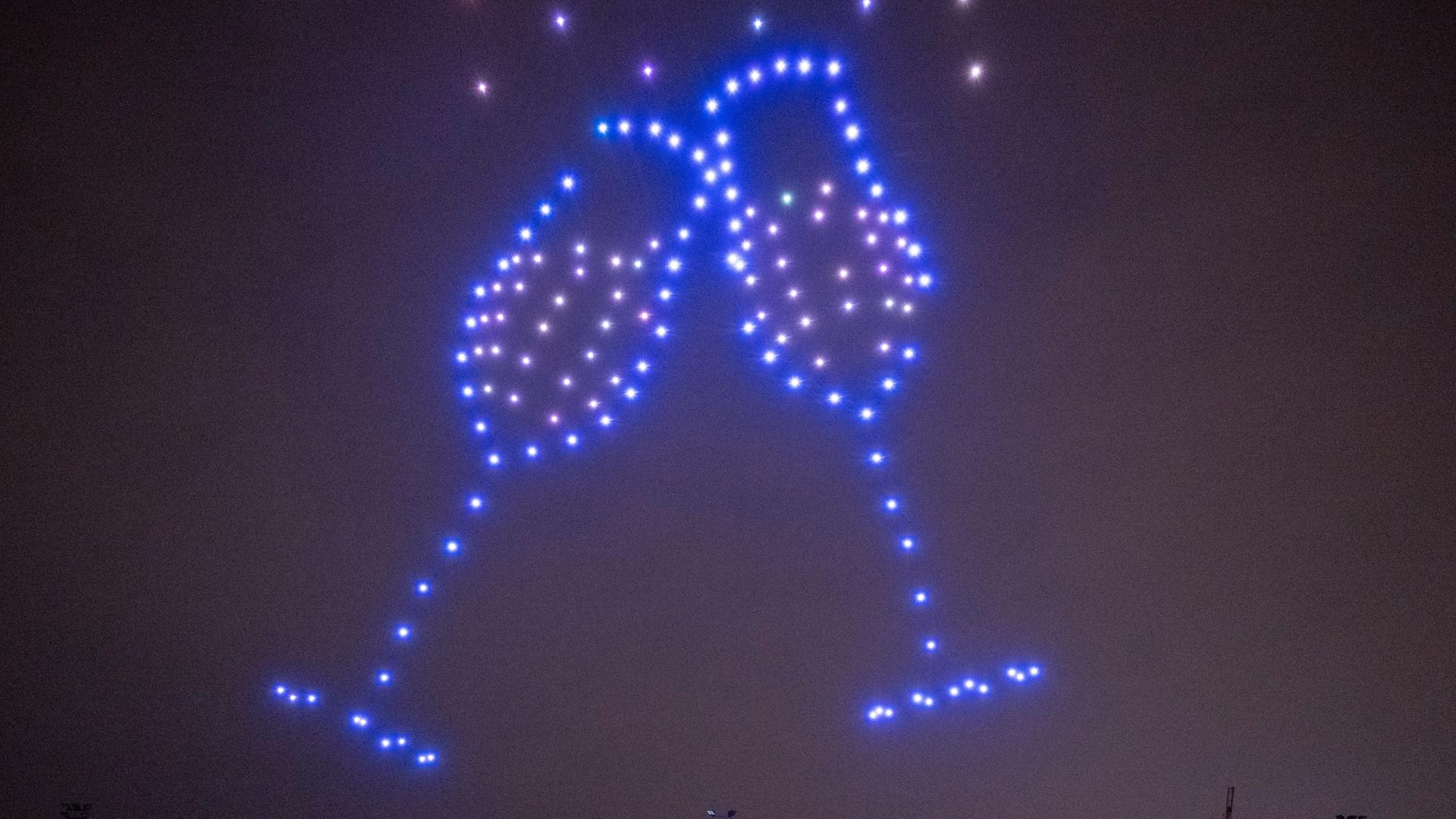 Leuchtende Drohnen fliegen bei einer Drohnenshow der Deutschen Umwelthilfe über einem Stadion und formen dabei am Himmel ein Bild von zwei Sektgläsern. 