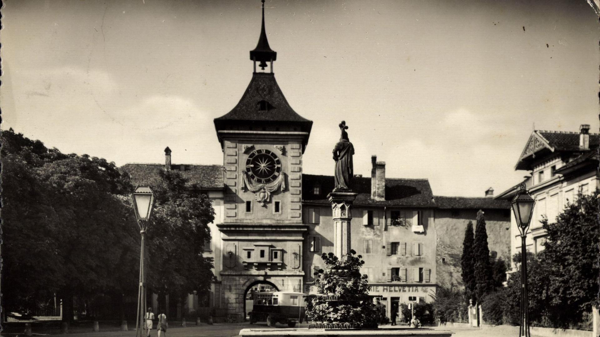 Ein historisches Schwarz-Weiß-Foto aus den 1930er-Jahren zeigt das Stadtzentrum von Murten mit Uhrturm, Denkmal und Laternen.
