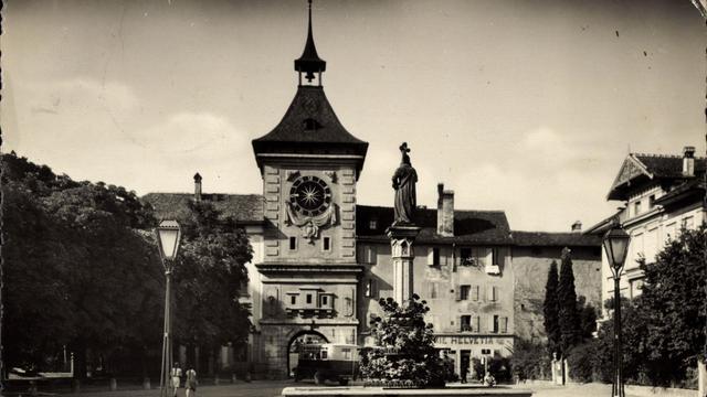 Ein historisches Schwarz-Weiß-Foto aus den 1930er-Jahren zeigt das Stadtzentrum von Murten mit Uhrturm, Denkmal und Laternen.
