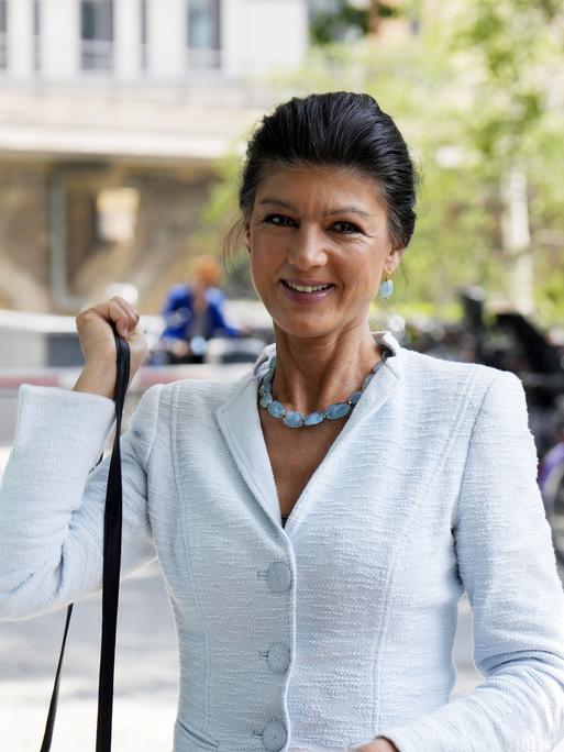 Sahra Wagenknecht lächelt in die Kamera und hängt sich dabei eine Handtasche an die Schulter. Im Hintergrund sieht man verschwommen abgestellte Fahrräder.