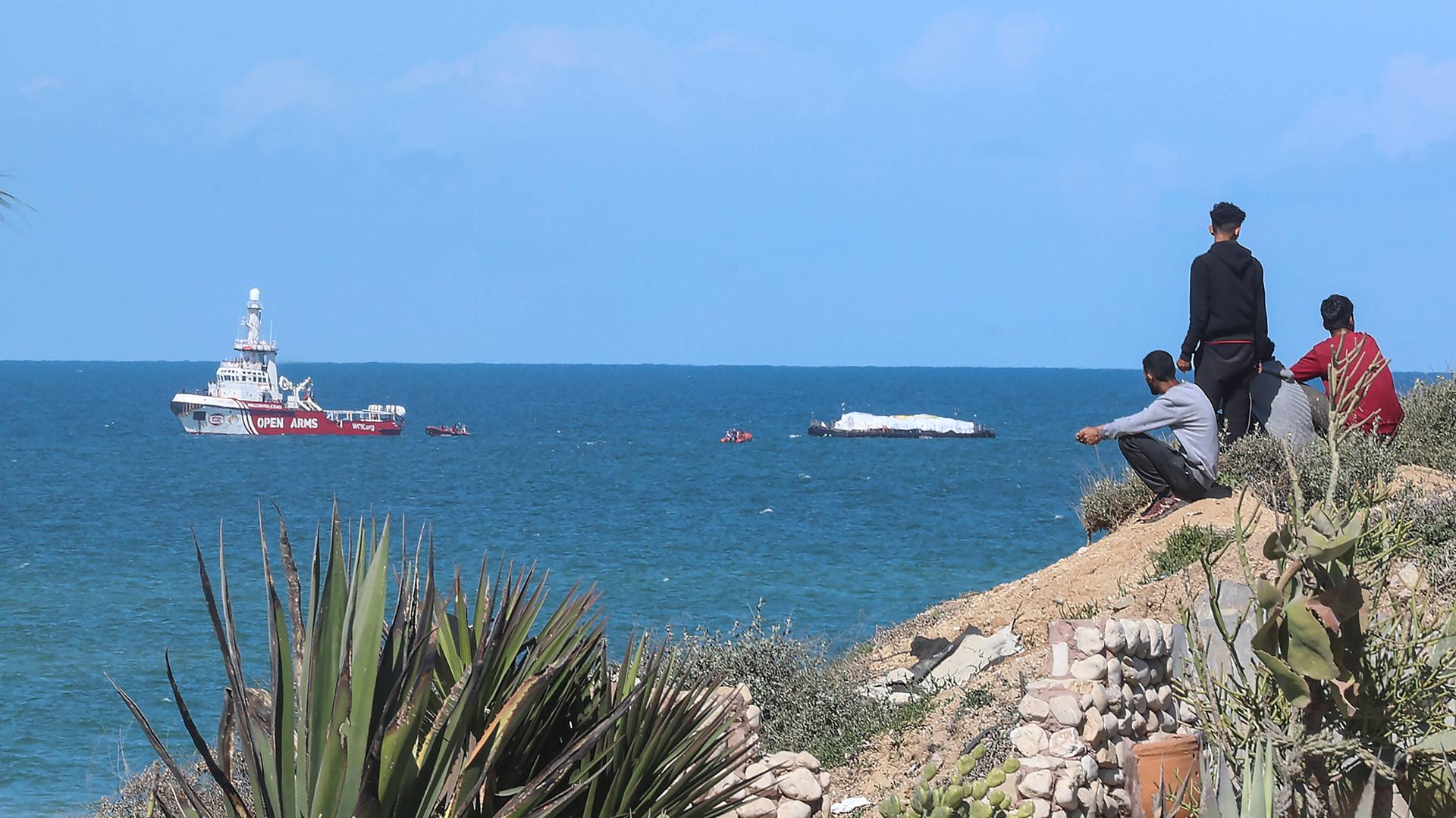 Mehrere Menschen beobachten an der Küste den Schlepper, der Hilfsgüter in den Gazastreifen bringt.