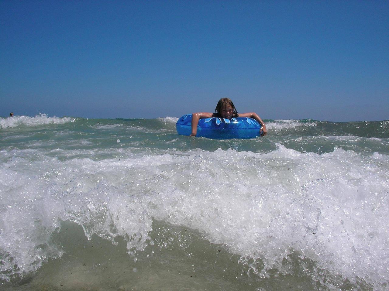 Ein Kind mit einem Schwimmreifen auf dem Meer. Im Vordergrund bricht gerade eine kleine Welle.