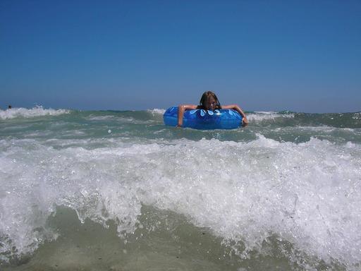 Ein Kind mit einem Schwimmreifen auf dem Meer. Im Vordergrund bricht gerade eine kleine Welle.