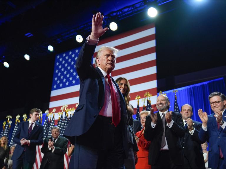 Donald Trump winkt. Hinter ihm stehen seine Frau und andere Menschen. 