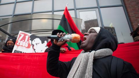 Mouctar Bah steht mit einem Mikrofon in der Hand vor einem roten Banner. Dahinter weiter Aktivisten, die Oury Jalloh gedenken.