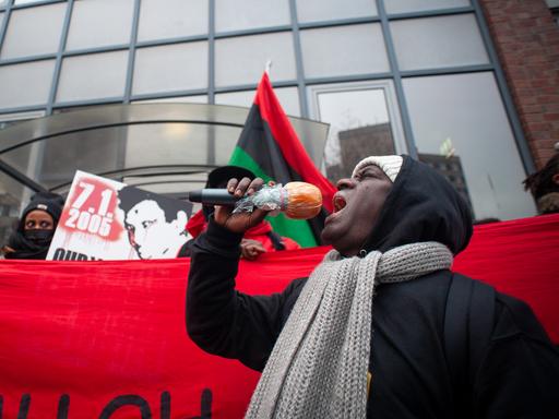 Mouctar Bah steht mit einem Mikrofon in der Hand vor einem roten Banner. Dahinter weiter Aktivisten, die Oury Jalloh gedenken.