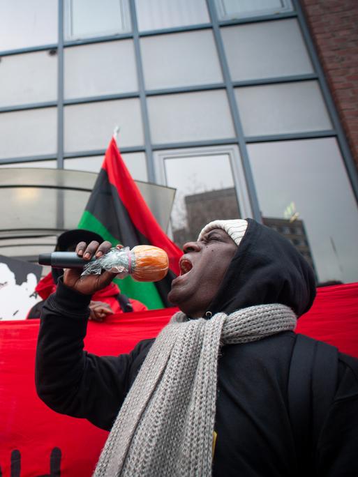 Mouctar Bah steht mit einem Mikrofon in der Hand vor einem roten Banner. Dahinter weiter Aktivisten, die Oury Jalloh gedenken.