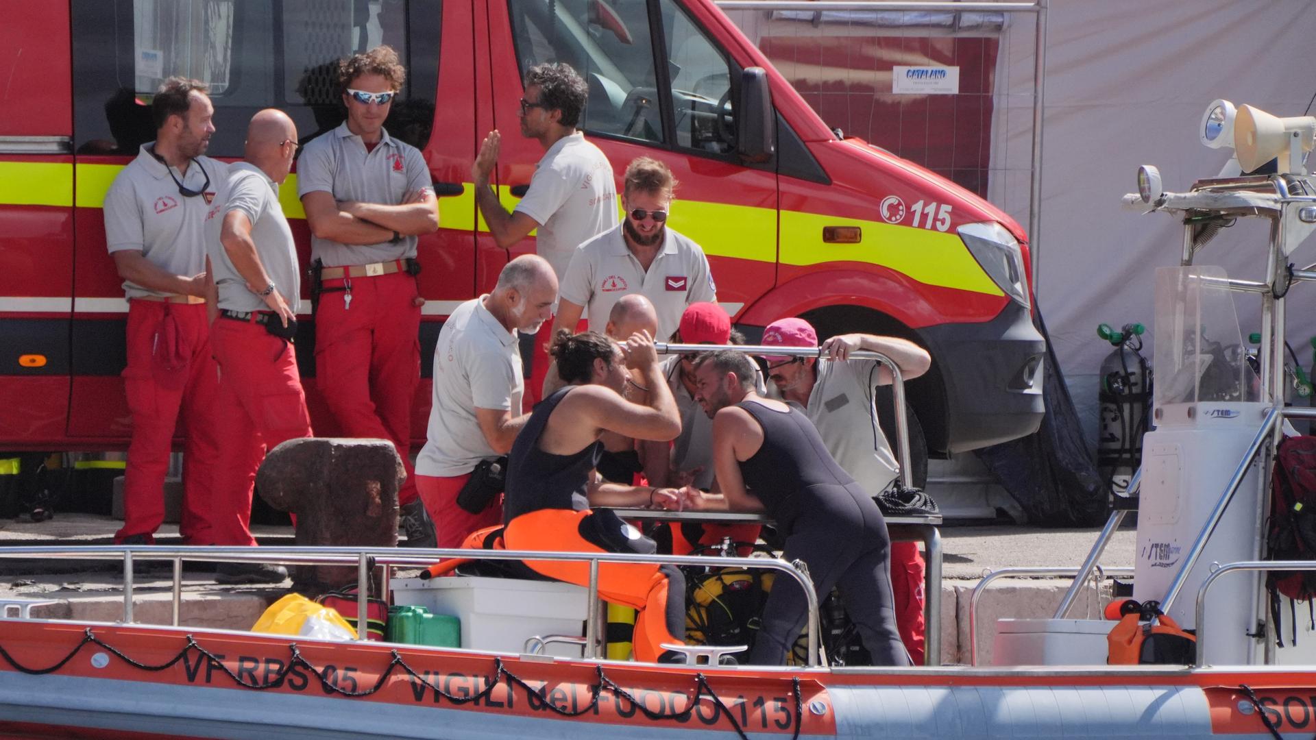 Zwei Taucher stehen auf einem kleinen Boot am Kai und unterhalten sich mit anderen Männern der Feuerwehr. Auf dem Kai steht ein Rettungsfahrzeug. 