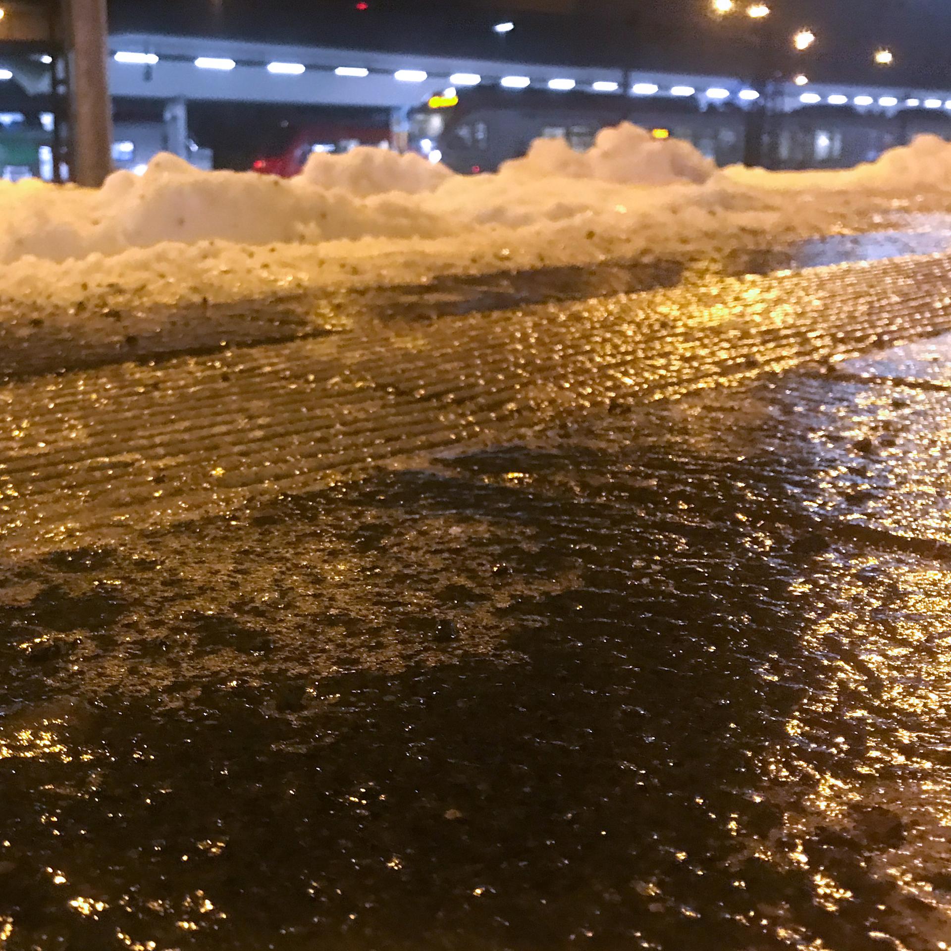 Blick auf eine spiegelglatte Straße mit Schnee am Rand; es ist dunkel. Im Hintergrund ein beleuchtetes Gebäude.