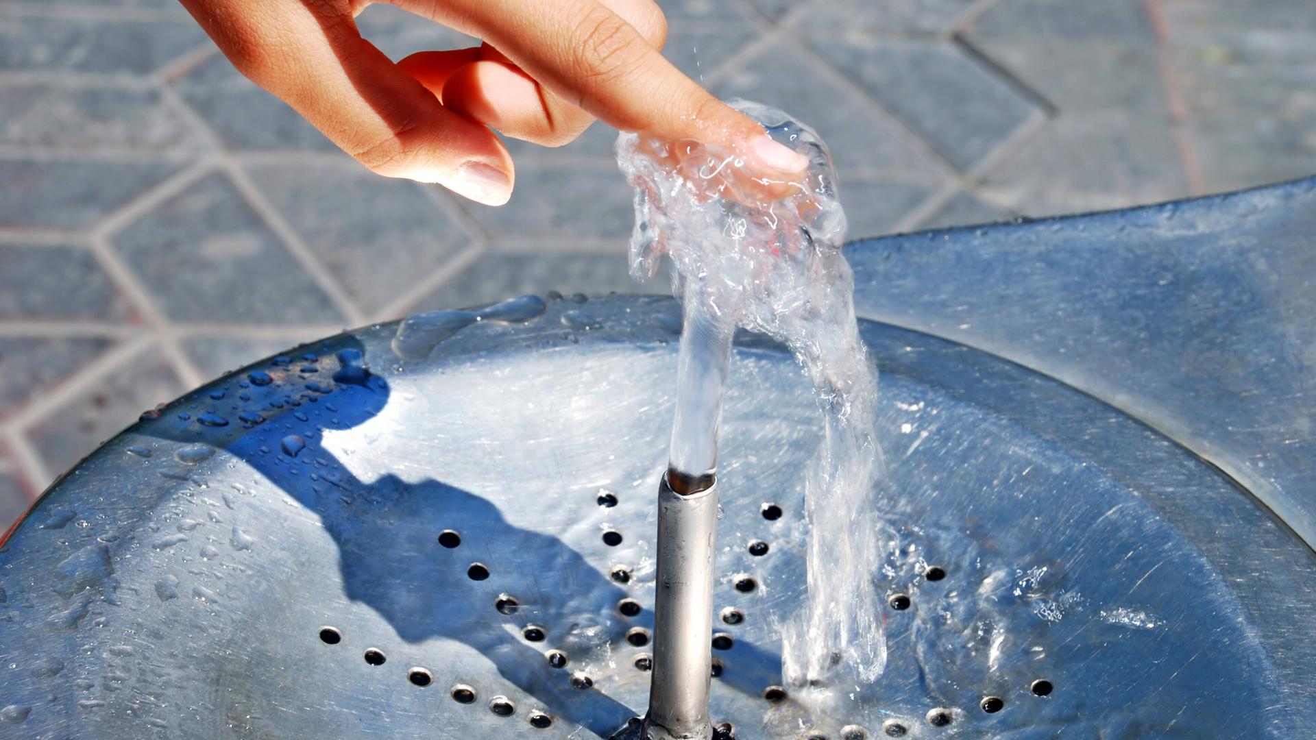 Eine Frau hält ihren Finger auf den Wasserstrahl eines Trinkwasserbrunnens.