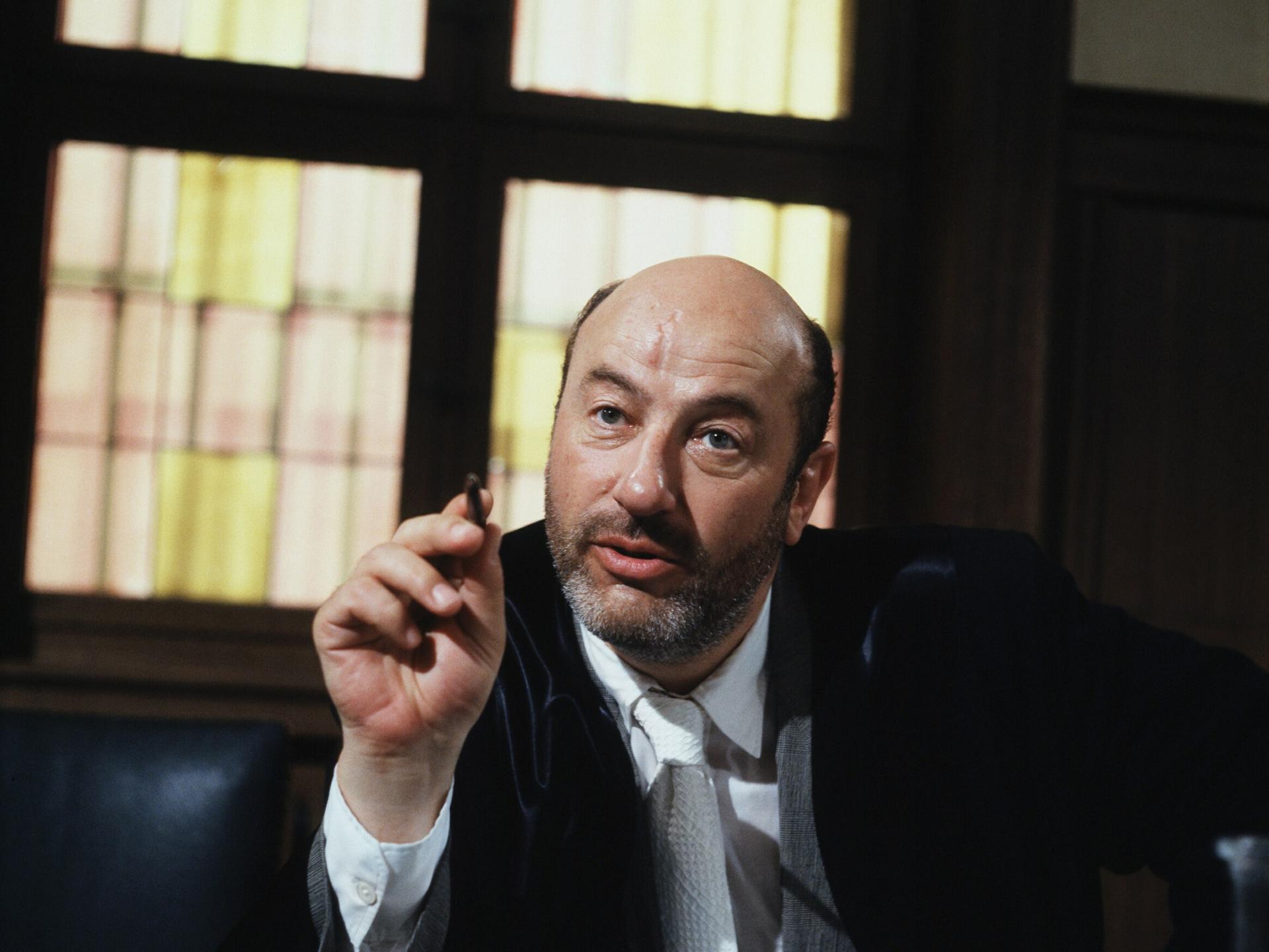 Manfred Krug in der Rolle des Anwalts Robert Liebling in der TV-Serie Liebling Kreuzberg. Er sitzt hinter einem massiven Schreibtisch und zeigt mit einem Stift in der rechten Hand nach vorn.
