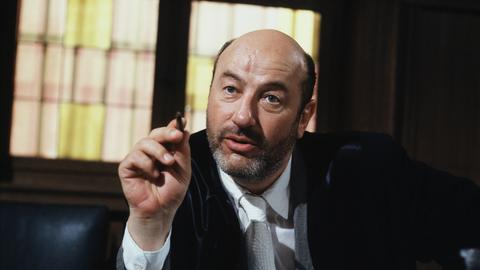 Manfred Krug in der Rolle des Anwalts Robert Liebling in der TV-Serie Liebling Kreuzberg. Er sitzt hinter einem massiven Schreibtisch und zeigt mit einem Stift in der rechten Hand nach vorn.