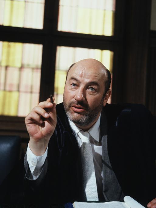 Manfred Krug in der Rolle des Anwalts Robert Liebling in der TV-Serie Liebling Kreuzberg. Er sitzt hinter einem massiven Schreibtisch und zeigt mit einem Stift in der rechten Hand nach vorn.