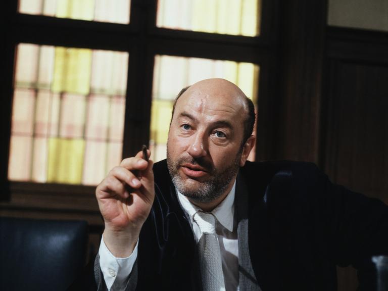 Manfred Krug in der Rolle des Anwalts Robert Liebling in der TV-Serie Liebling Kreuzberg. Er sitzt hinter einem massiven Schreibtisch und zeigt mit einem Stift in der rechten Hand nach vorn.