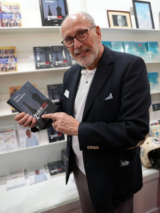 Jörg H. Trauboth lacht in die Kamera. Er steht in einer Buchhandlung und hält sein eigenes Buch "Zarentod" in der Hand.
