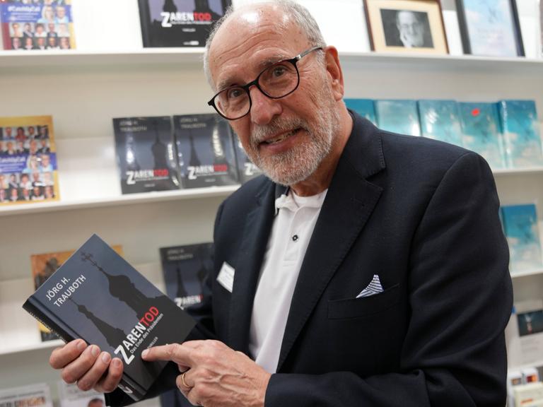 Jörg H. Trauboth lacht in die Kamera. Er steht in einer Buchhandlung und hält sein eigenes Buch "Zarentod" in der Hand.