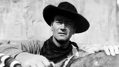 John Wayne trägt einen Cowboyhut und legt die Hand auf den Sattel seines Pferdes.