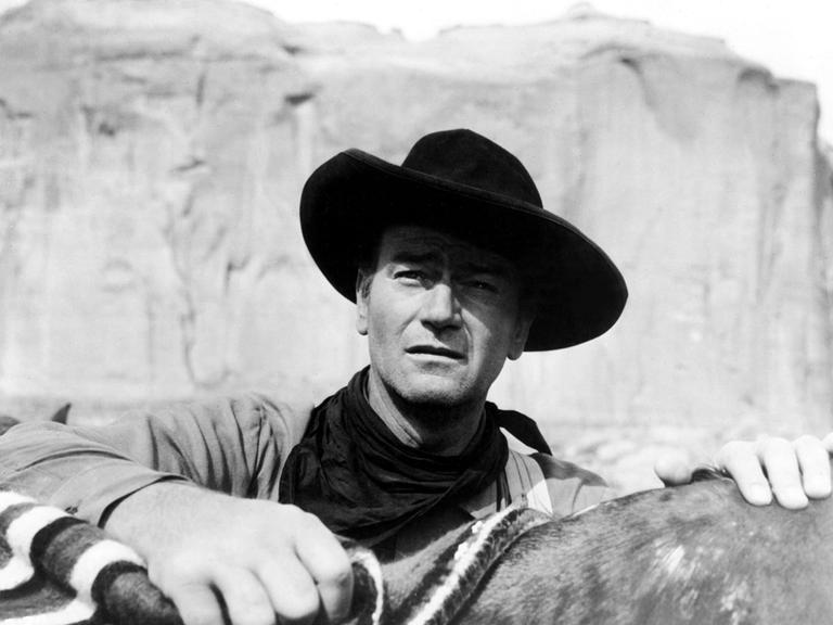 John Wayne trägt einen Cowboyhut und legt die Hand auf den Sattel seines Pferdes.