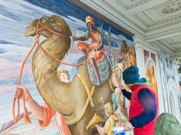 Rekonstruktionsarbeiten an einem Bild im Dresdener Residenzschloss. Eine Frau malt an einem Wandbild, das ein Kamel mit Reiter zeigt.