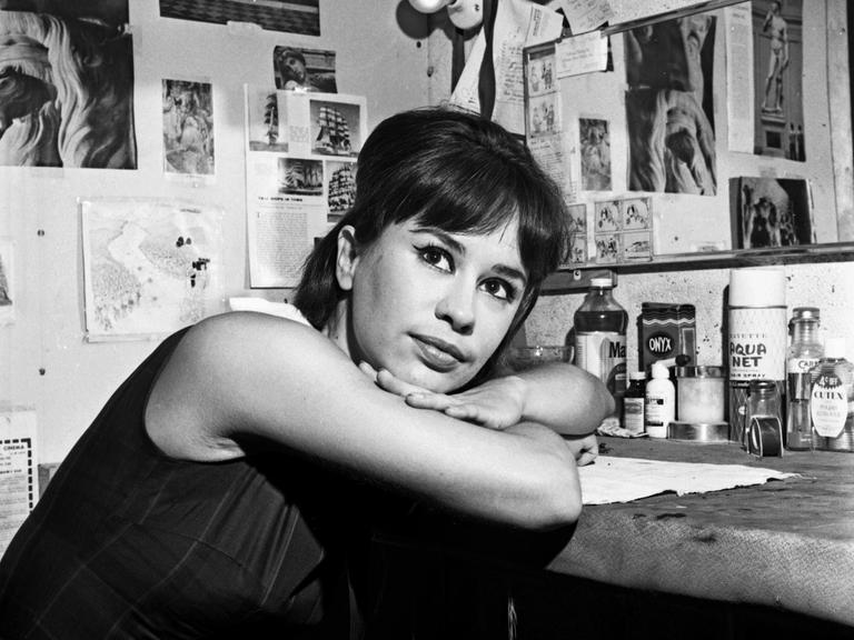 Astrud Gilberto lehnt auf einem Tisch auf dem viele Gegenstände stehen und schaut in die Kamera in diesem schwarz-weiss Foto.