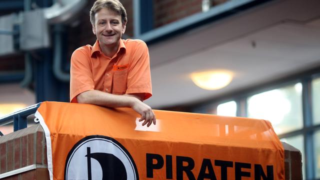Jens Seipenbusch blickt auf dem Bundesparteitag der Piratenpartei in Hamburg in die Kamera des Fotografen. Er trägt ein orangefarbenes Hemd, vor ihm hängt eine Fahne der Piratenpartei am Geländer des Treppenhauses.