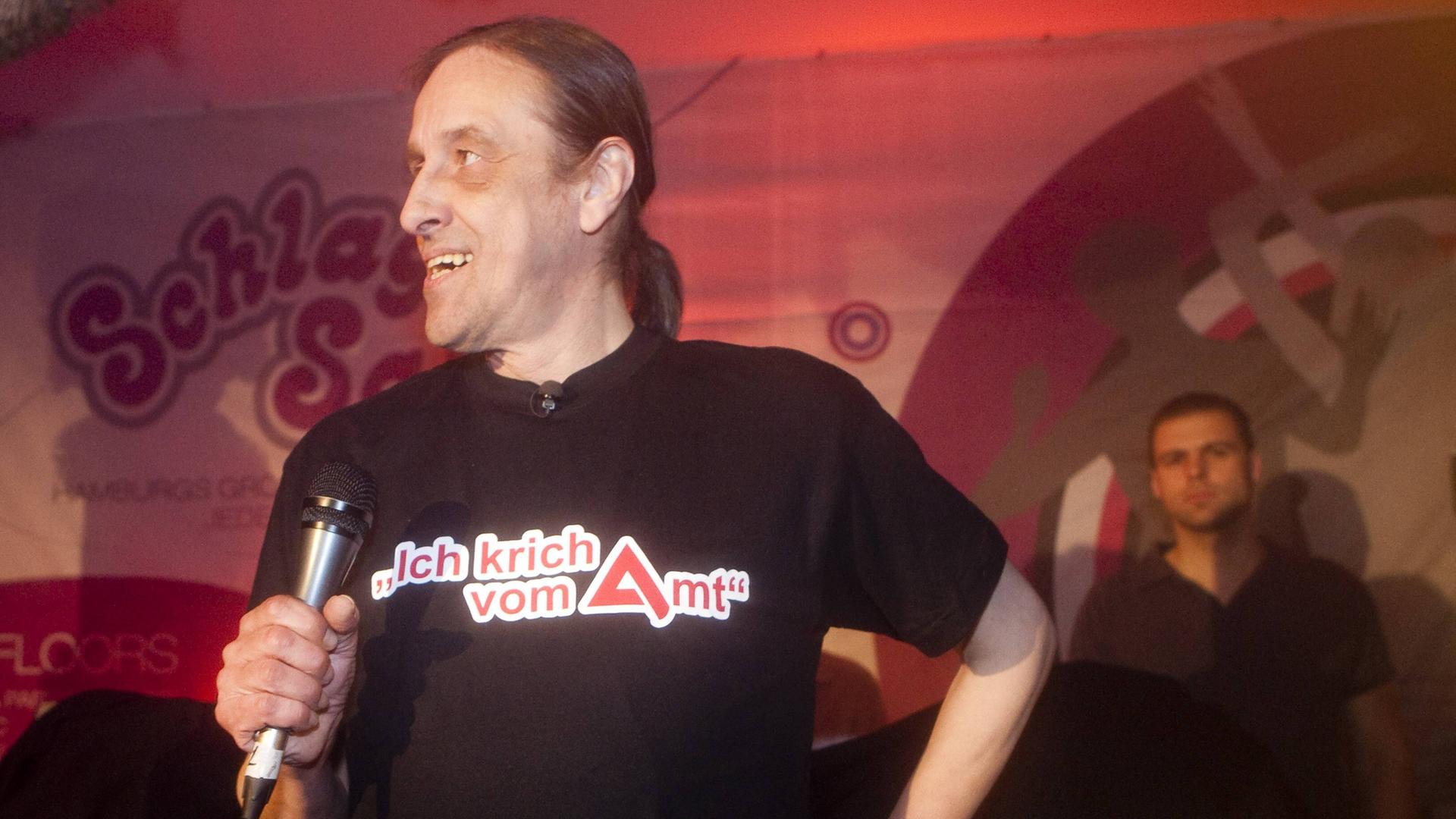 Arno Dübel steht mit einem Mikrofon in der Hand auf der Bühne. Auf seinem T-Shirt steht "Ich krich vom Amt".