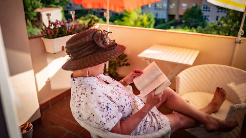 Ein Frau mit Sonnenhut sitzt auf dem Balkon und liest ein Buch.