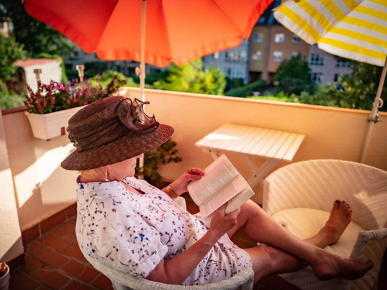 Ein Frau mit Sonnenhut sitzt auf dem Balkon und liest ein Buch.