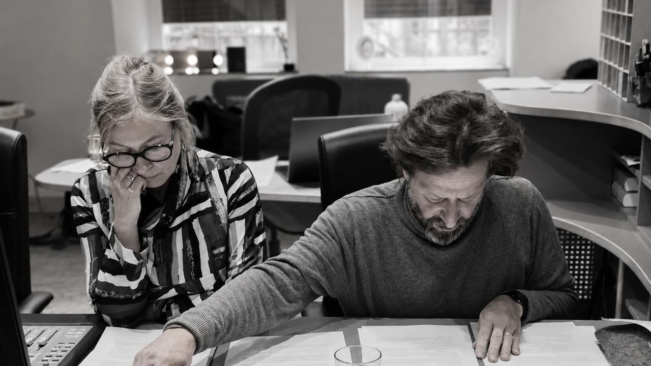 Zwei Personen (das Autorenteam Hannah Hofmann und Sven Lindholm)sitzen an einem Schreibtisch und prüfen Dokumente; links ein Mischpult mit Reglern, in der Mitte ein Glas Wasser, im Hintergrund Büroarbeitsplätze und Regale. Zwei Personen (das Autorenteam Hannah Hofmann und Sven Lindholm)sitzen an einem Schreibtisch und prüfen Dokumente; links ein Mischpult mit Reglern, in der Mitte ein Glas Wasser, im Hintergrund Büroarbeitsplätze und Regale.