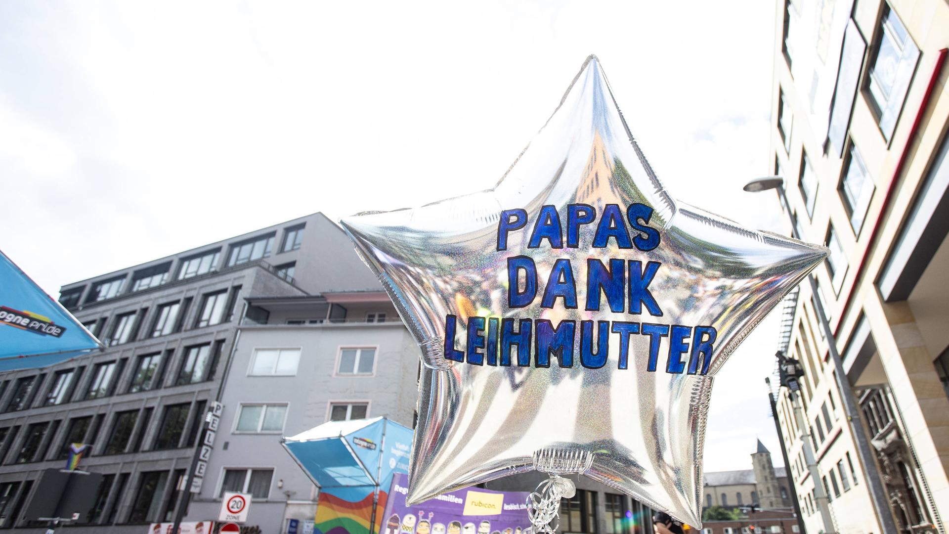 Eine CSD-Parade in Köln. Dabei wird ein silberner Luftballon in Sternform in die Höhe gehalten. Darauf steht "Papas, dank Leihmutter". Eine CSD-Parade in Köln. Dabei wird ein silberner Luftballon in Sternform in die Höhe gehalten. Darauf steht "Papas, dank Leihmutter".