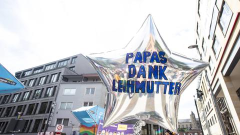 Eine CSD-Parade in Köln. Dabei wird ein silberner Luftballon in Sternform in die Höhe gehalten. Darauf steht "Papas, dank Leihmutter".