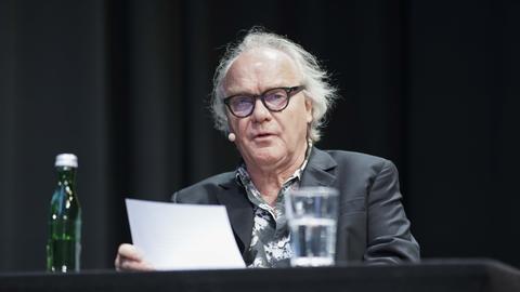 Ein Mann mit grauem Haar und Brille sitzt auf eine Bühne und liest vor. 