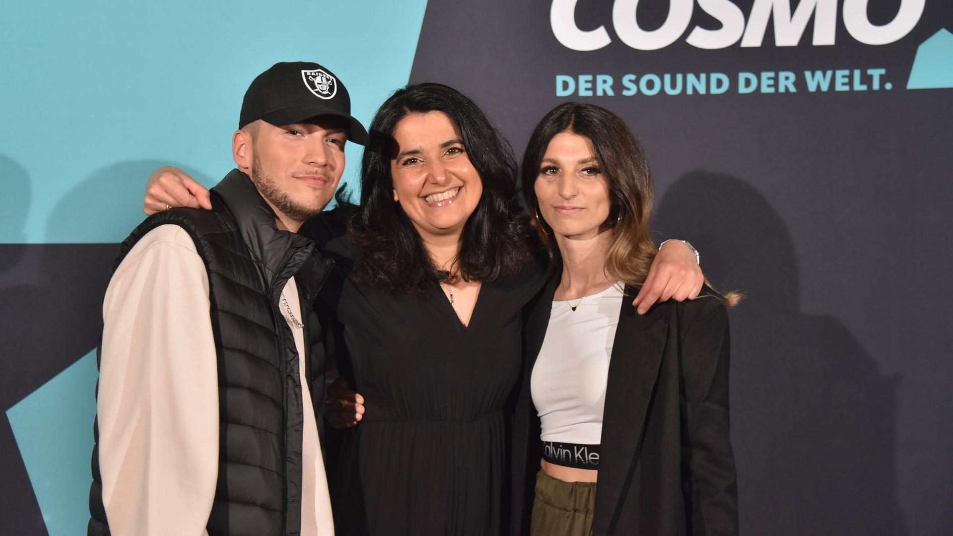 Cosmo-Redakteurin Schiwa Schlei (Mitte) mit Autor Patrick Phul (links) und Regisseurin Esra Phul. Sie stehen auf vor einer Fotowand zur Premiere der Rap-Musical-Serie "Hype", im Hintergrund rechts ist das Cosmo-Logo zu sehen. 