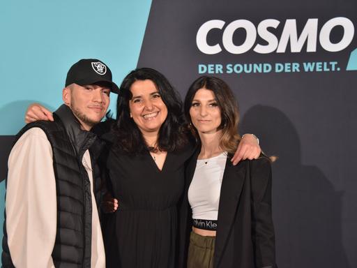 Cosmo-Redakteurin Schiwa Schlei (Mitte) mit Autor Patrick Phul (links) und Regisseurin Esra Phul. Sie stehen auf vor einer Fotowand zur Premiere der Rap-Musical-Serie "Hype", im Hintergrund rechts ist das Cosmo-Logo zu sehen. 