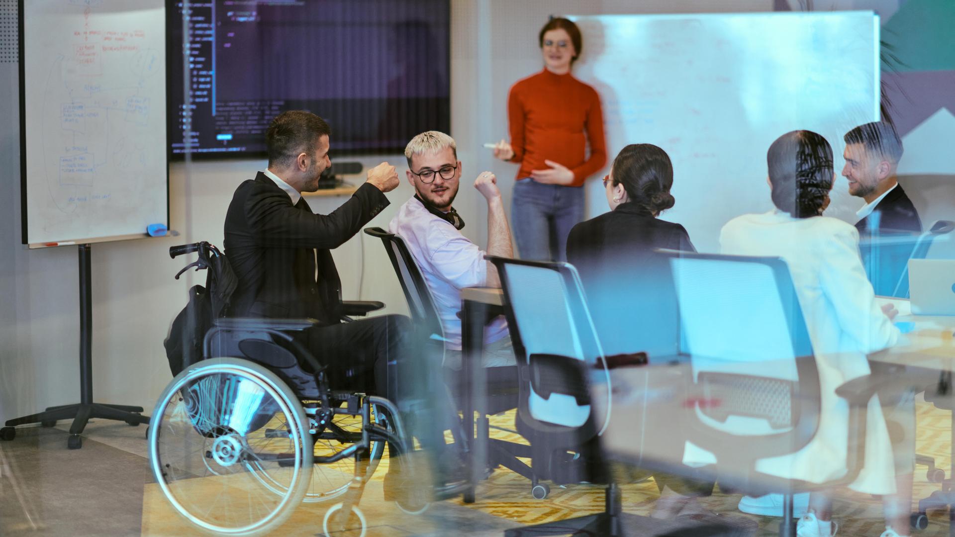 Eine schwangere Geschäftsfrau präsentiert ihren Kollegen in einem modernen Glasbüro ihren Geschäftsplan. Ein Mitarbeiter im Anzug sitzt in einem Rollstuhl neben dem Gruppentisch. Eine schwangere Geschäftsfrau präsentiert ihren Kollegen in einem modernen Glasbüro ihren Geschäftsplan. Ein Mitarbeiter im Anzug sitzt in einem Rollstuhl neben dem Gruppentisch.