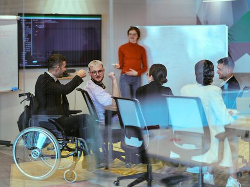 Eine schwangere Geschäftsfrau präsentiert ihren Kollegen in einem modernen Glasbüro ihren Geschäftsplan. Ein Mitarbeiter im Anzug sitzt in einem Rollstuhl neben dem Gruppentisch. Eine schwangere Geschäftsfrau präsentiert ihren Kollegen in einem modernen Glasbüro ihren Geschäftsplan. Ein Mitarbeiter im Anzug sitzt in einem Rollstuhl neben dem Gruppentisch.