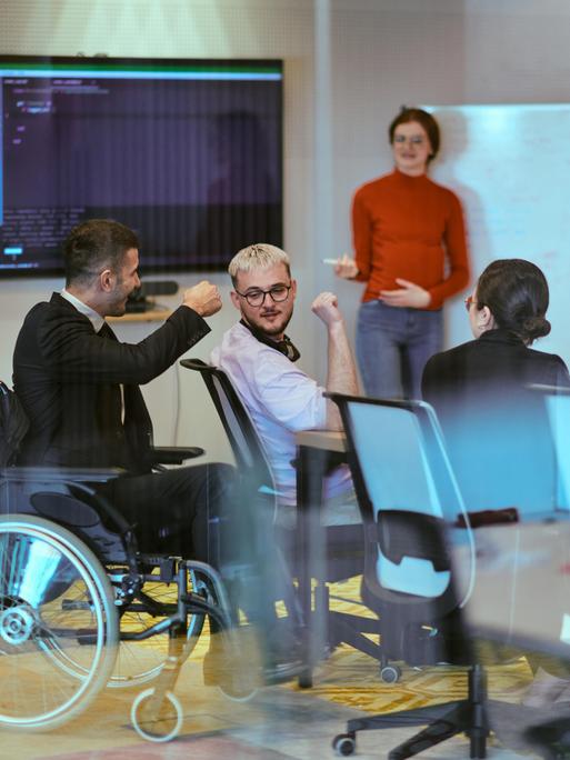 Eine schwangere Geschäftsfrau präsentiert ihren Kollegen in einem modernen Glasbüro ihren Geschäftsplan. Ein Mitarbeiter im Anzug sitzt in einem Rollstuhl neben dem Gruppentisch. Eine schwangere Geschäftsfrau präsentiert ihren Kollegen in einem modernen Glasbüro ihren Geschäftsplan. Ein Mitarbeiter im Anzug sitzt in einem Rollstuhl neben dem Gruppentisch.