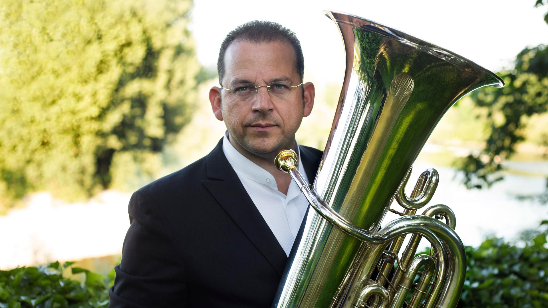 Ein Mann mit kurzen dunklen Haaren posiert mit Tuba im Grünen. Er trägt Brille, ein weißes Hemd und ein schwarzes Sakko.