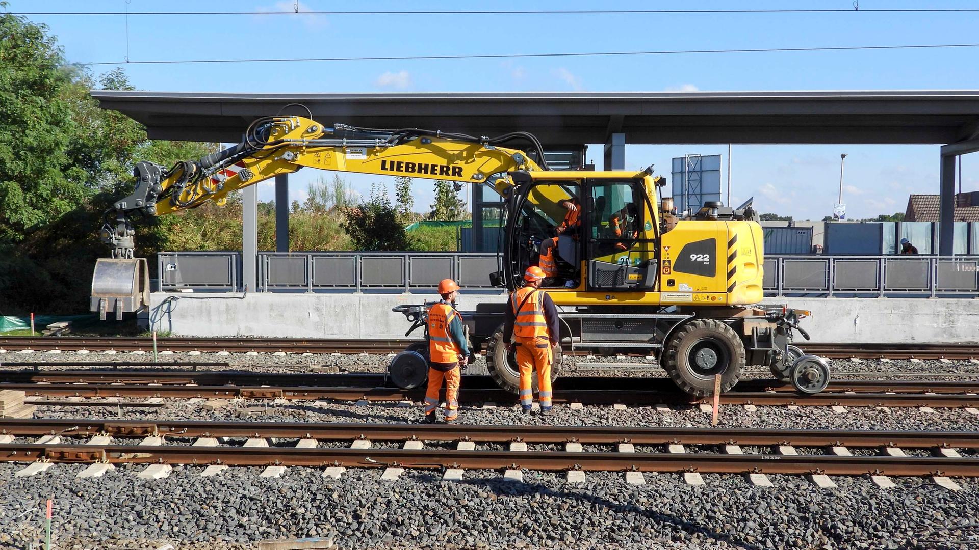 Zwei Arbeiter in Schutz-Kleidung stehen vor einem Bahnhof. Hinter ihnen ist ein Bagger auf Schienen.
