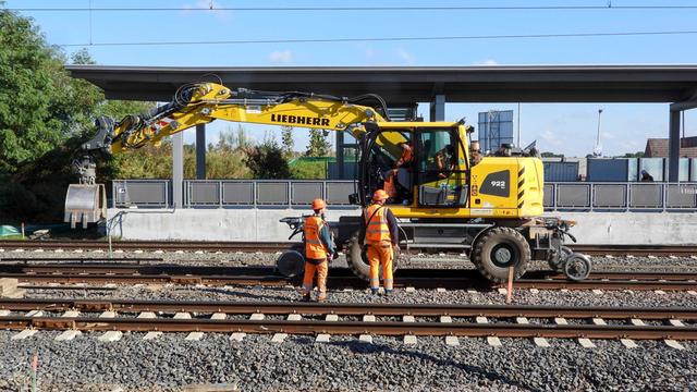 Zwei Arbeiter in Schutz-Kleidung stehen vor einem Bahnhof. Hinter ihnen ist ein Bagger auf Schienen.