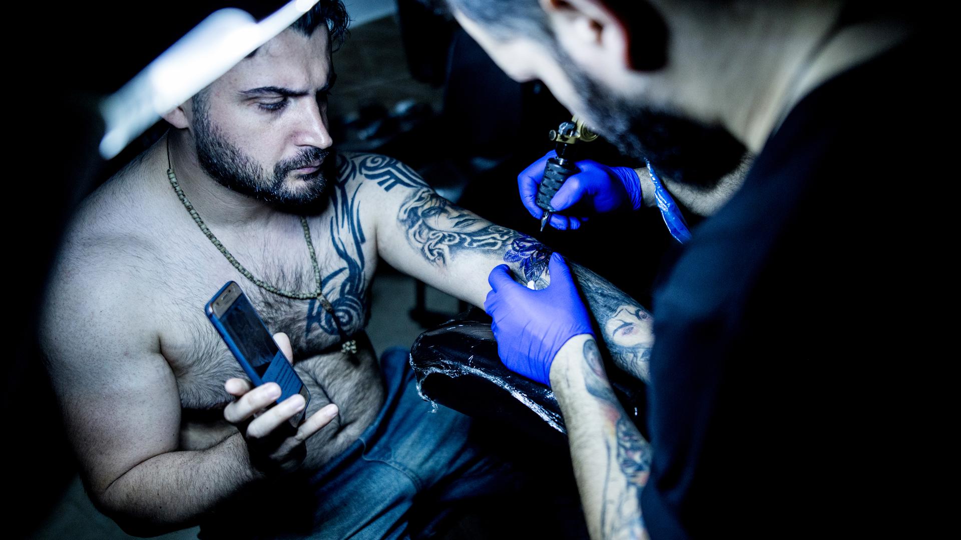 Ein iranischer Tätowierer in seinem Tattoostudio bei der Arbeit, er tätowiert einen Mann dessen Haut bei einem Unfall verbrannt wurde, Gilan im Iran, 2019.