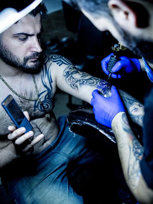 Ein iranischer Tätowierer in seinem Tattoostudio bei der Arbeit, er tätowiert einen Mann dessen Haut bei einem Unfall verbrannt wurde, Gilan im Iran, 2019.